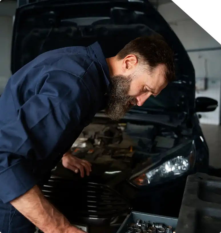 Een monteur met een baard in een blauw uniform onderzoekt met een geconcentreerde blik een automotor. De motorkap is open, waardoor de motoronderdelen in een garage zichtbaar zijn.